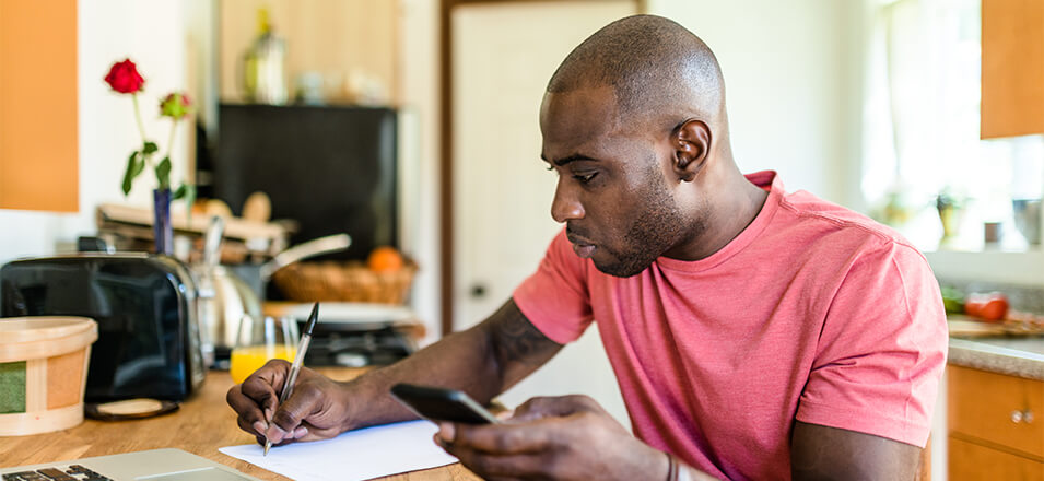 African American man sitting at kitchen counter making a plan with his laptop, phone, and pad of paper